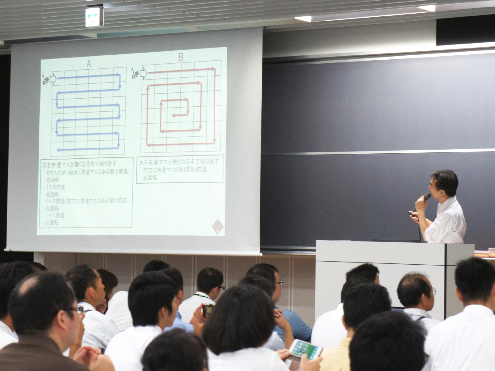 ロボット掃除機が全マスを通るルートを2通り示し、それぞれプログラミングで実現するとどんな特徴やメリットがあるのかを説明する鷲崎氏