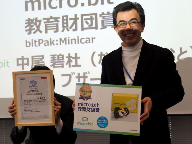 課題解決部門のmicro:bit教育財団賞を受賞した中尾碧杜さんは当日参加できなかったが（写真は代理）、越塚教授からbitPark Minicarが贈られた