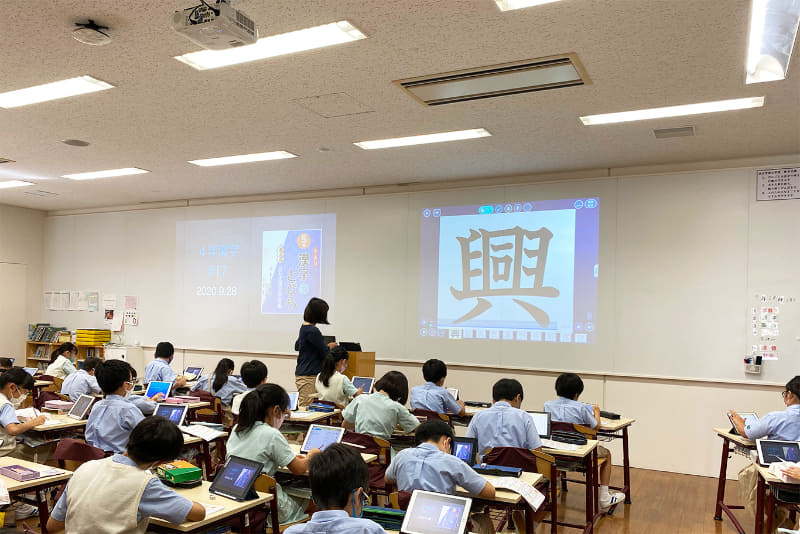 授業風景。校内はWi-Fi完備。iPad活用を進めていくうちに、教員から「黒板で2つ違うものを見せたい」というリクエストが寄せられるように。全普通教室の横壁をホワイトボードにして、プロジェクターを各教室に2台ずつ整備。もともと黒板前に設置されていたものと合わせて、各普通教室には計3台のプロジェクターが整備されている