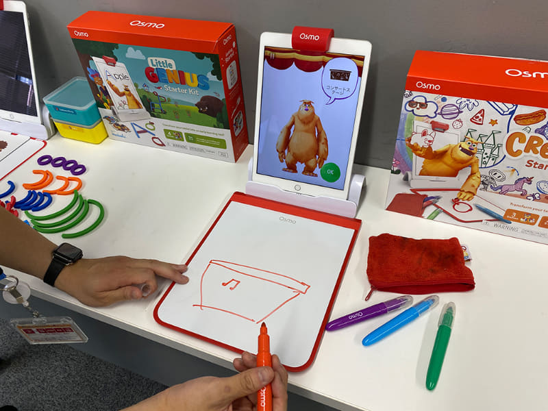 「Monster」のゲームで、Moから「コンサートステージ」のお題を出され挑戦する筆者。「Osmo クリエイティブ スターター キット」には、何度も消せるクリエイティブボードと4色のドライイレースマーカー、文字を消すときにも使えるイレイザーポーチが付属している