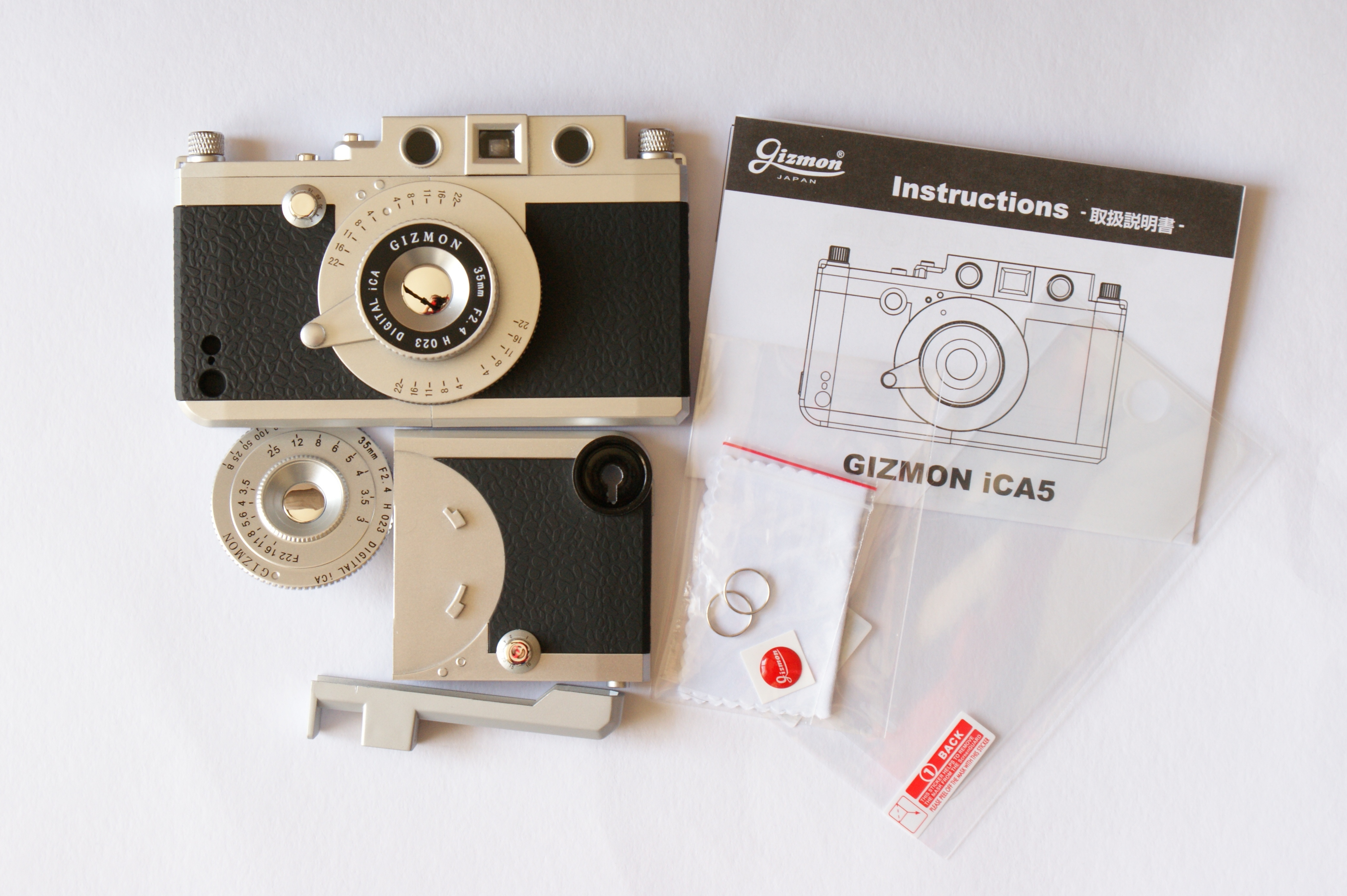 同梱物は（左上から時計回りに）ケース本体、説明書、液晶保護フィルム、ロゴ入りホームボタンシール・クリーニングクロス・ストラップ取付用のリング、別売のフィルターやコンバージョンレンズ取付用のケース、三脚取付用アダプタ、薄型のモックレンズ